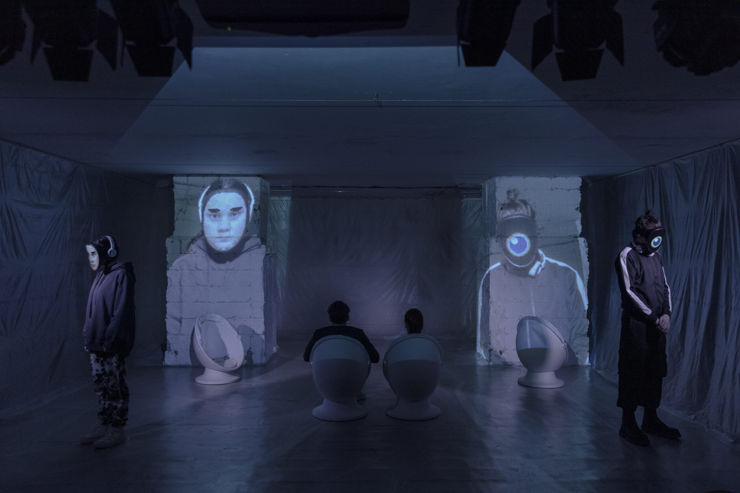 Temná miestnosť s projekciami tvárí na stene, dvaja ľudia sedia na bielych stoličkách, dvaja stoja so svetelnými maskami.