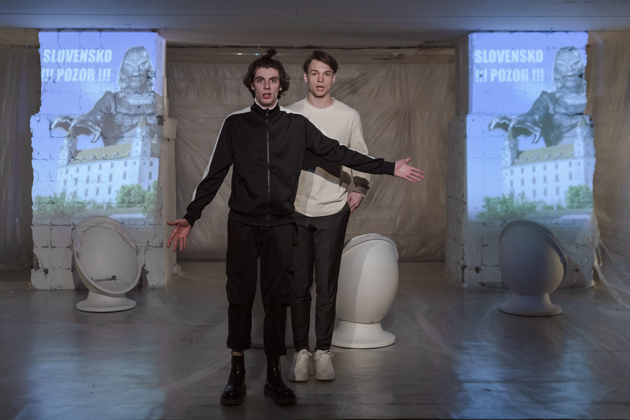 Dvaja muži stoja pred premietanou scénou s nápisom „SLOVENSKO POZOR!“ a obrazom Bratislavského hradu s obrovským monštrom.