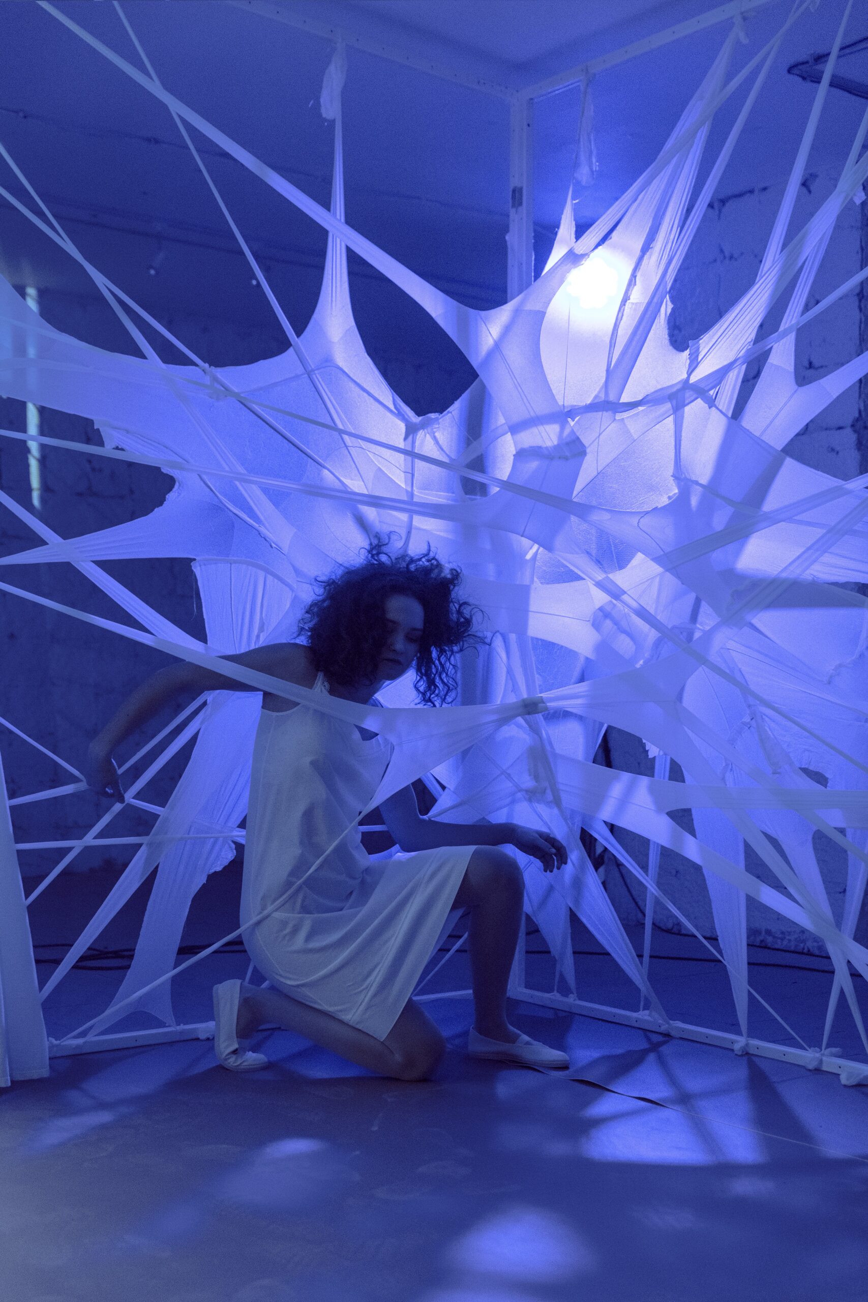 Mladá žena v bielych šatách uprostred scény vytvorenej z bielych pások látky napodobňujúcich pavučinu, so surrealistickým modrým osvetlením.