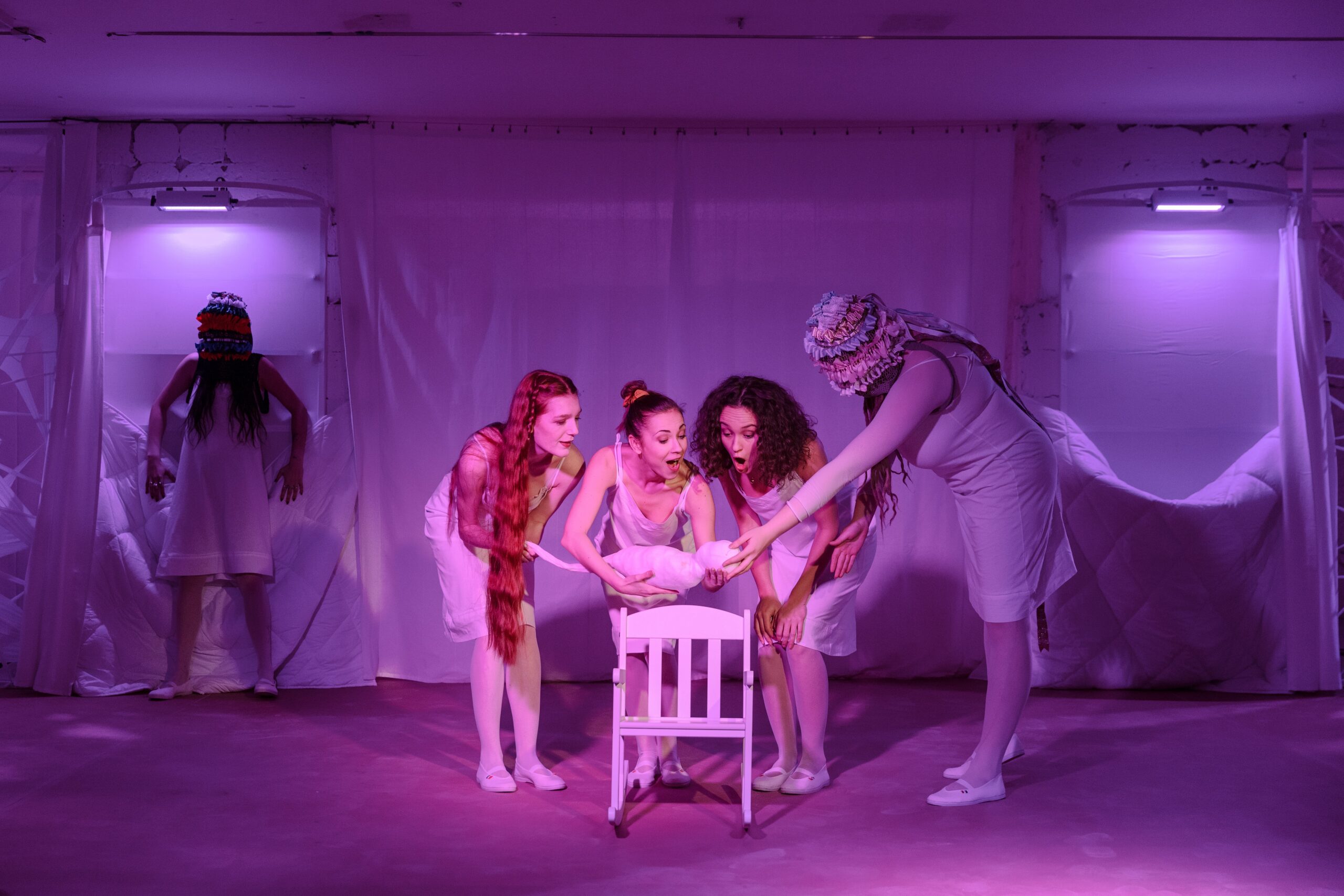 Na scéne skupina štyroch mladých žien v bielych šatách. Radostne sa skláňajú nad prázdnu, bielu stoličku, v rukách držia dieťa. V pozadí stojí postava. Fialové osvetlenie.