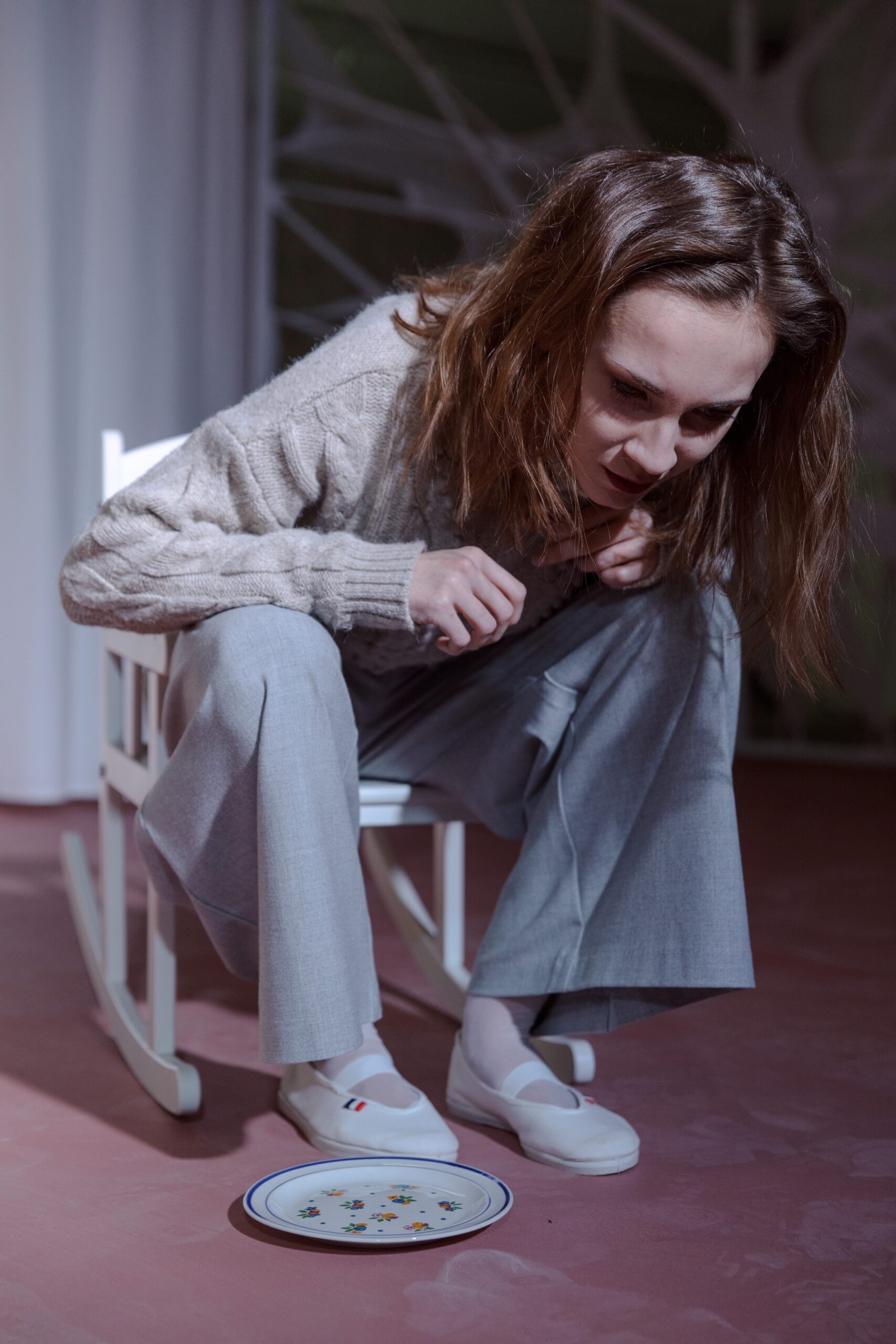 Žena v pletenom svetri a sivých nohaviciach sedí na bielej stoličke, skláňa sa nad prázdny tanier s kvetinovým vzorom, ktorý leží na zemi.