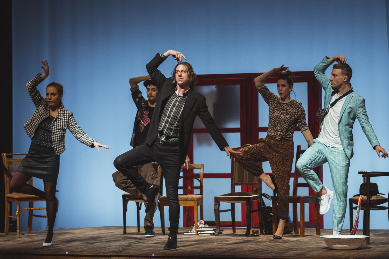 Päť ľudí v dynamických pózach na pódiu, v dramatickej scéne, s hnedými stoličkami v pozadí. Modré osvetlenie.