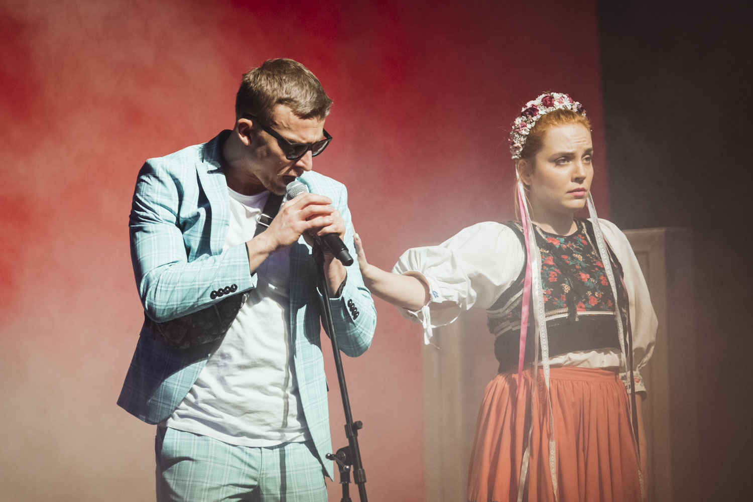 Muž v belasom kockovanom obleku spieva do mikrofónu, žena v tradičnom kroji stojí bokom s rozpaženou rukou, na pozadí červené dymové efekty.