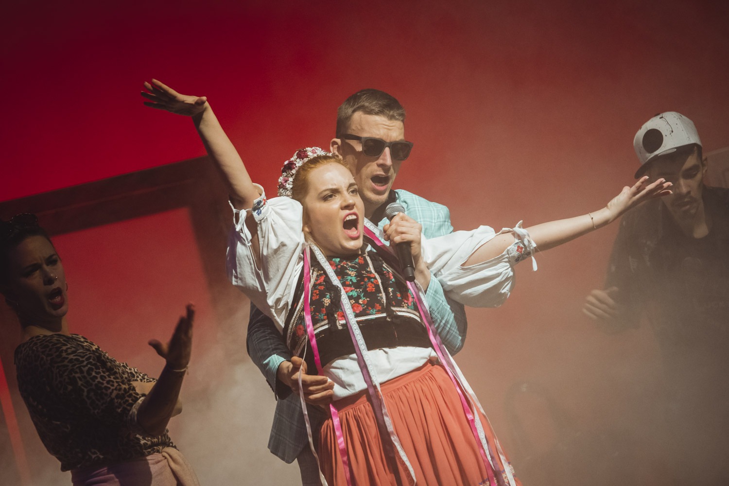 Skupina ľudí vo výrazných pózach spieva a gestikuluje na pódiu. V popredí žena v tradičnom kroji a muž v belasom obleku. V pozadí červená hmla.