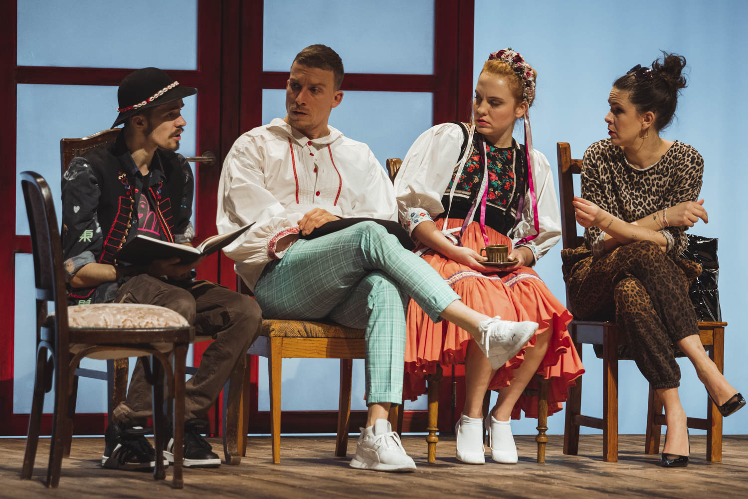 Štyri diskutujúce osoby sediace vedľa seba, zľava: muž s knihou, muž v kockovaných belasých nohaviciach, žena v ľudovom kroji a žena v leopardom odeve. Modré osvetlenie.