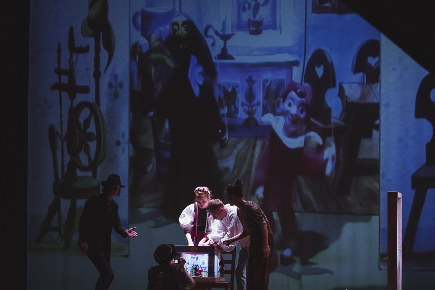 Celok. Scéna s piatimi hercami v rôznych kostýmoch, ktorí hrajú bábkové divadlo, v pozadí veľká projekcia s bábkami.