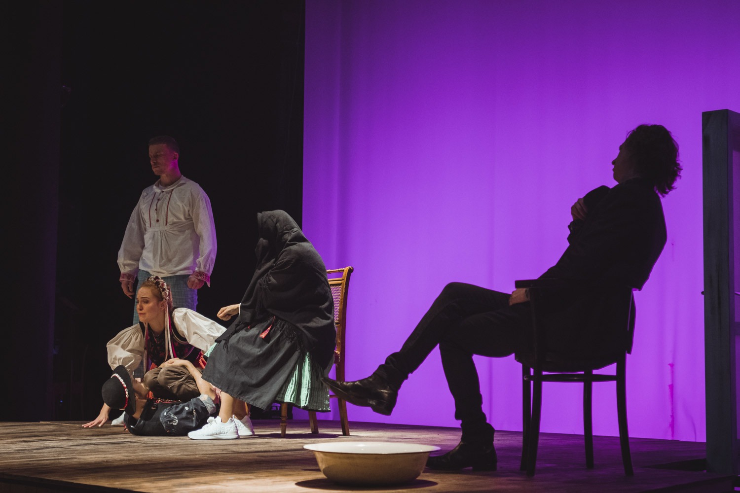 Postavy v tradičných krojoch v dramatickej póze na javisku. Muž stojaci nad dvoma ženami a mužom ležiacim na zemi. V popredí muž sediaci v tieni na stoličke. Fialové pozadie.