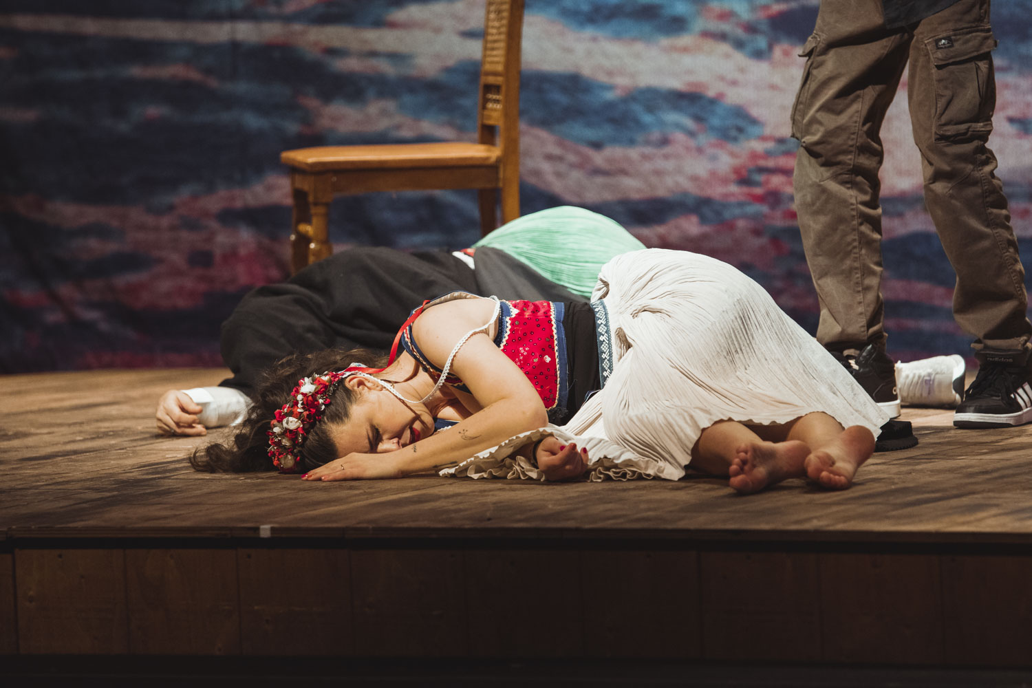 Dve ženy v ľudovom kroji ležiace na drevenej podlahe javiska, nad nimi stojí muž.
