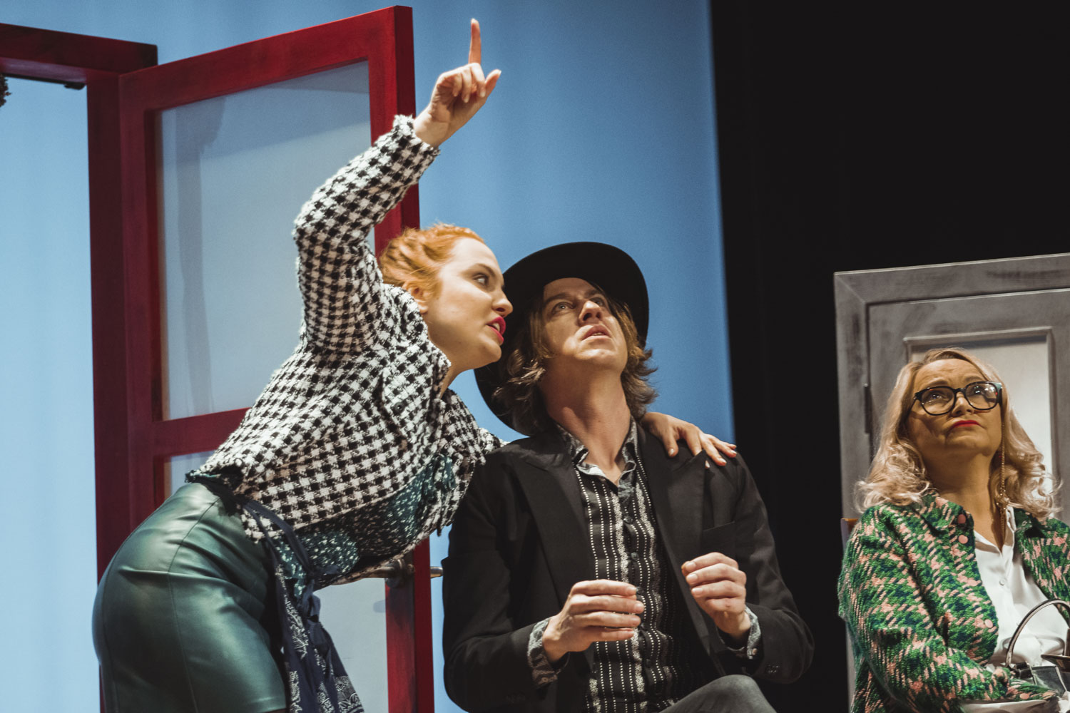 Dve osoby v interakcii, muž v čiernom klobúku sedí a hľadí nahor. Žena, ktorá je k nemu naklonená ukazuje prstom smerom nahor, v pozadí sedí žena v zeleno-čiernom károvanom saku.