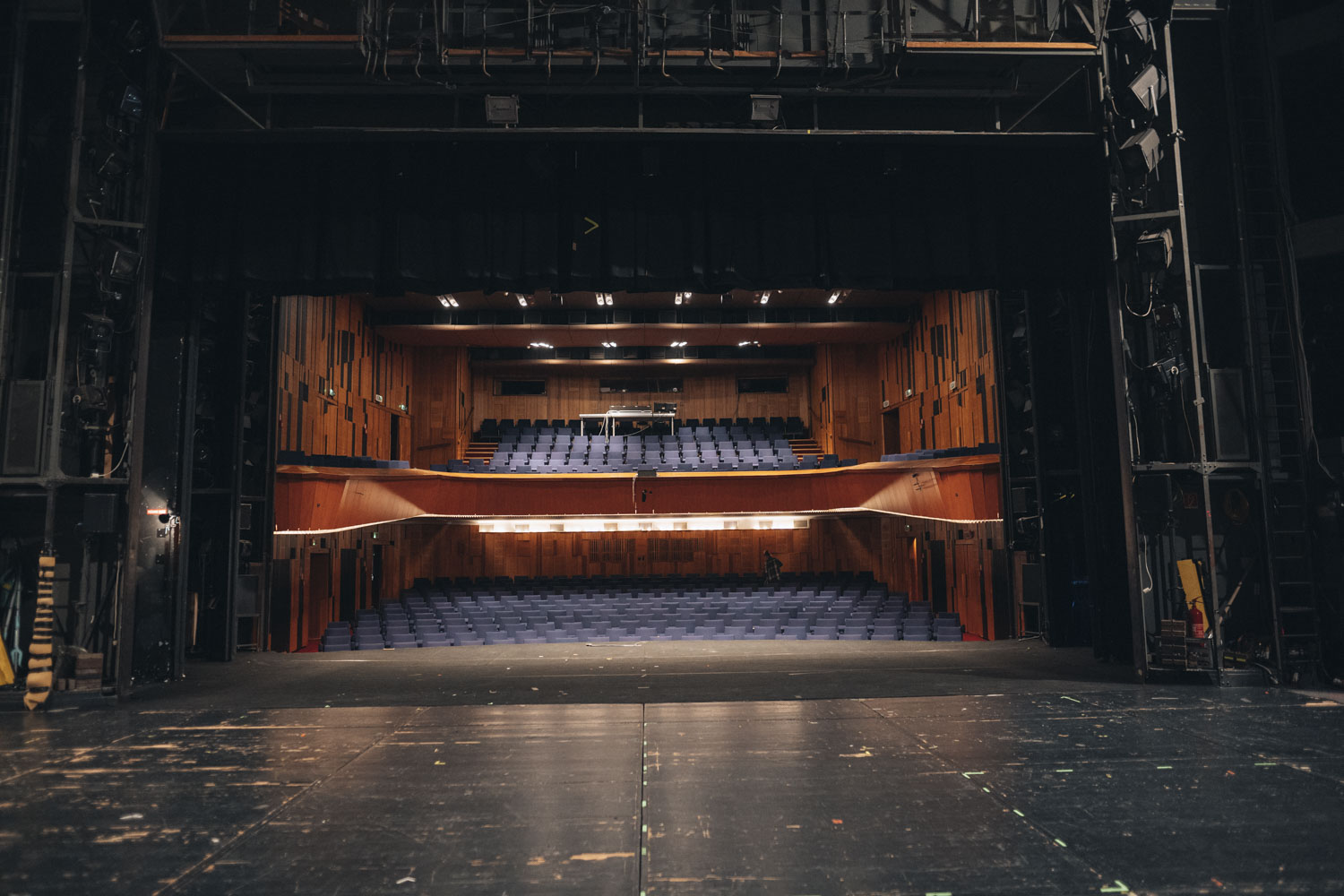 Interiér divadla. Pohľad zo stredu prázdneho javiska smerom do prázdneho hľadiska divadla s modrými sedadlami a dreveným obkladom na stenách.