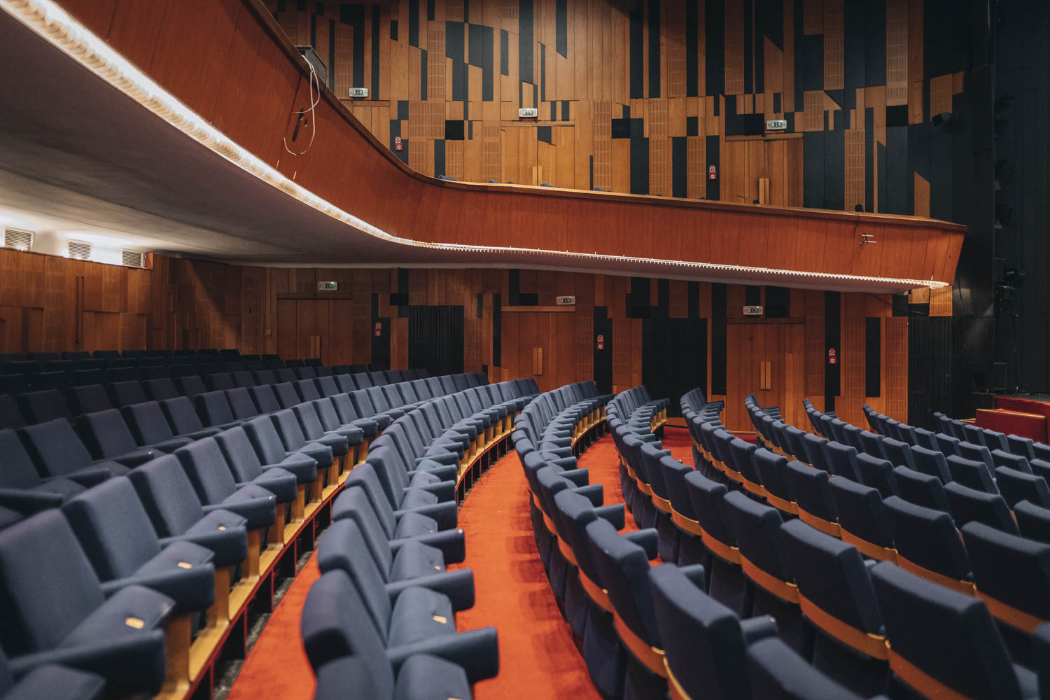 Divadelná sála. Jemné osvetlenie. Podhľad z pravej strany hľadiska na divadelný balkón, spoza modrých sedadiel hľadiska. Steny sály majú drevený obklad.