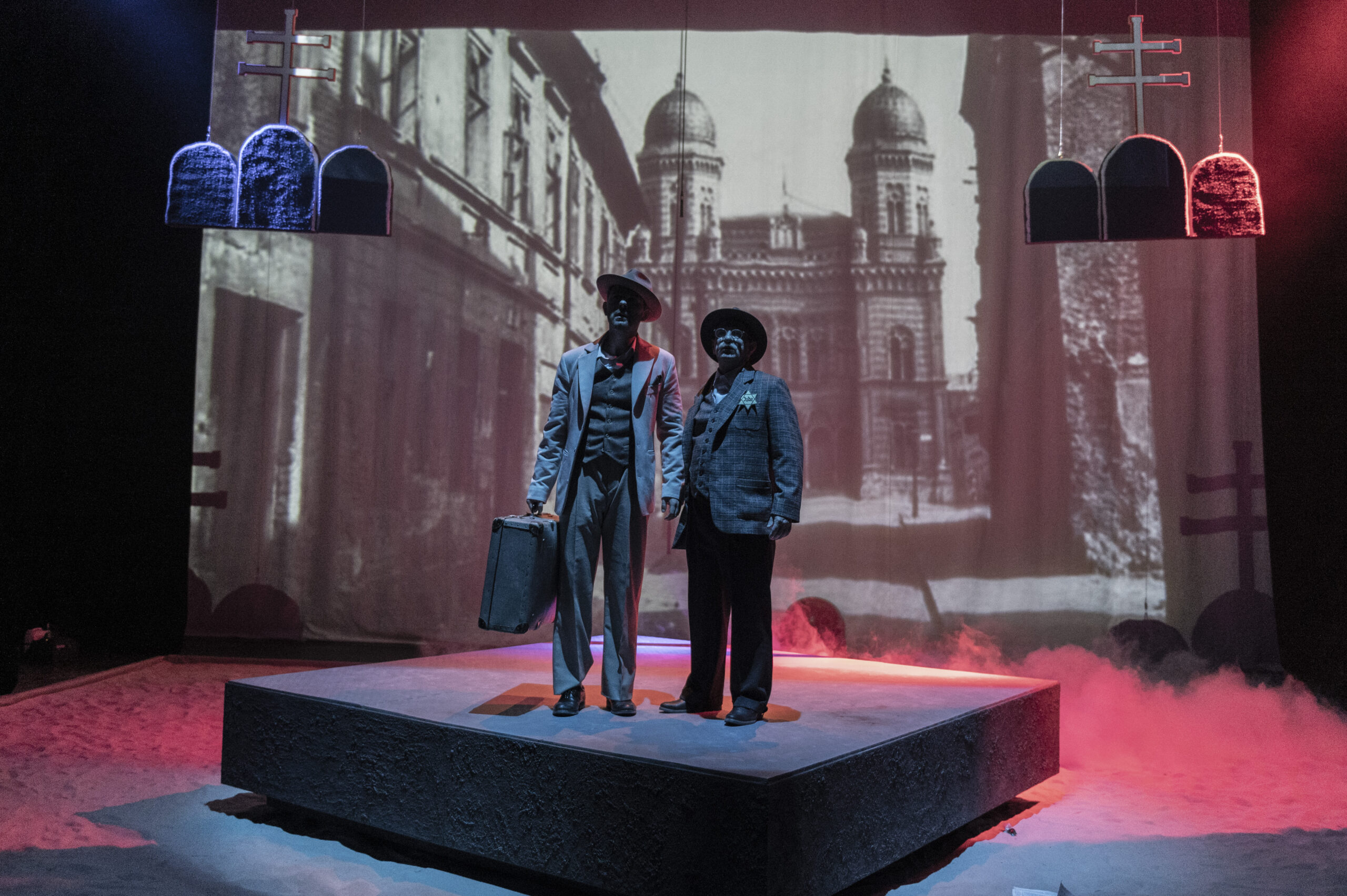 Dvaja muži v oblekoch s pripnutou Dávidovou hviezdou stoja na pódiu s fotografiou synagógy v pozadí.