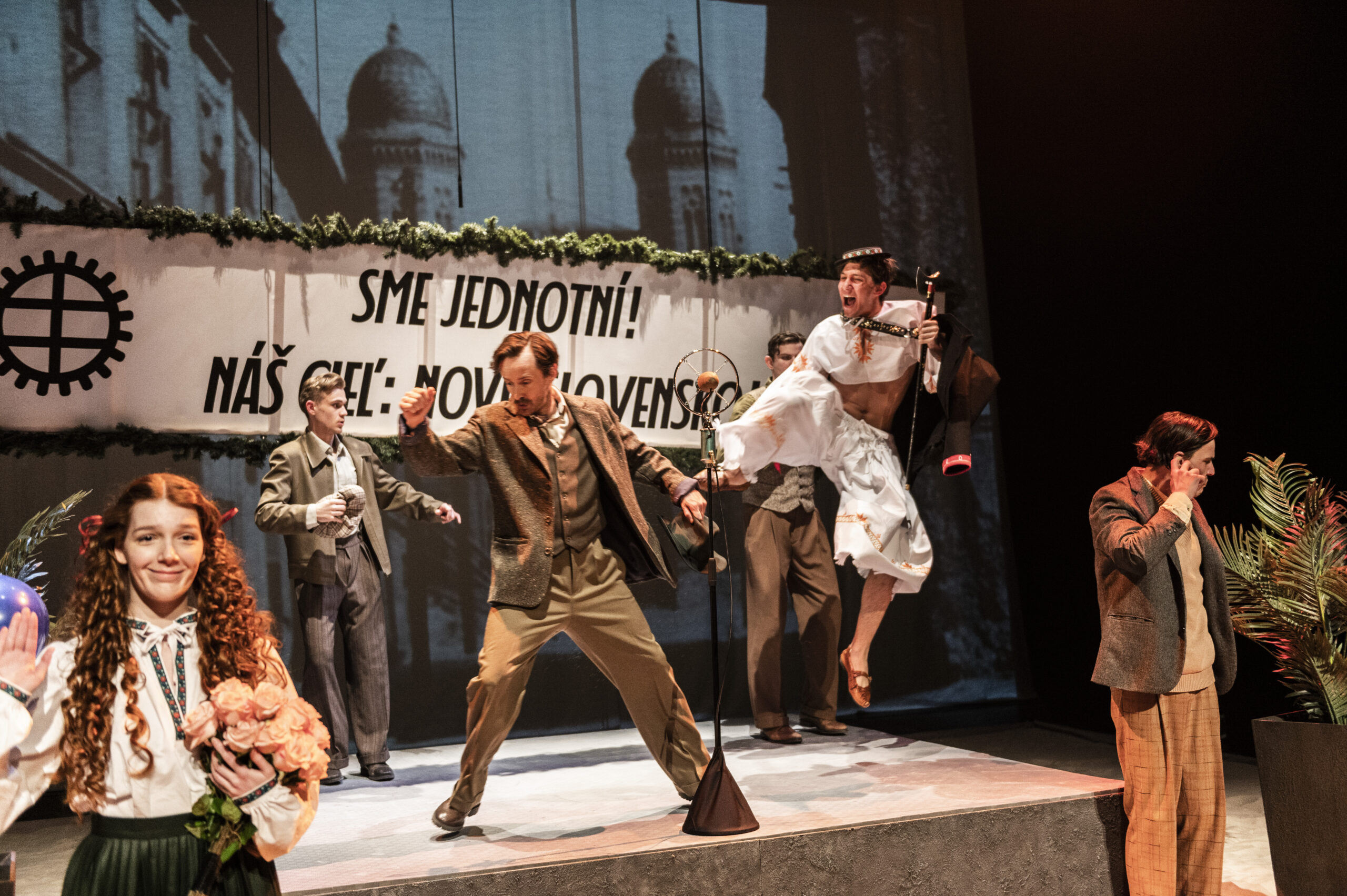 Živá javisková scéna s viacerými postavami v tradičných kostýmoch, jedna žena drží kvety, muž v detvianskom koostýme vyskakuje držiac valašku. V pozadí je nápis Sme jednotní! Náš cieľ: Nové Slovensko!