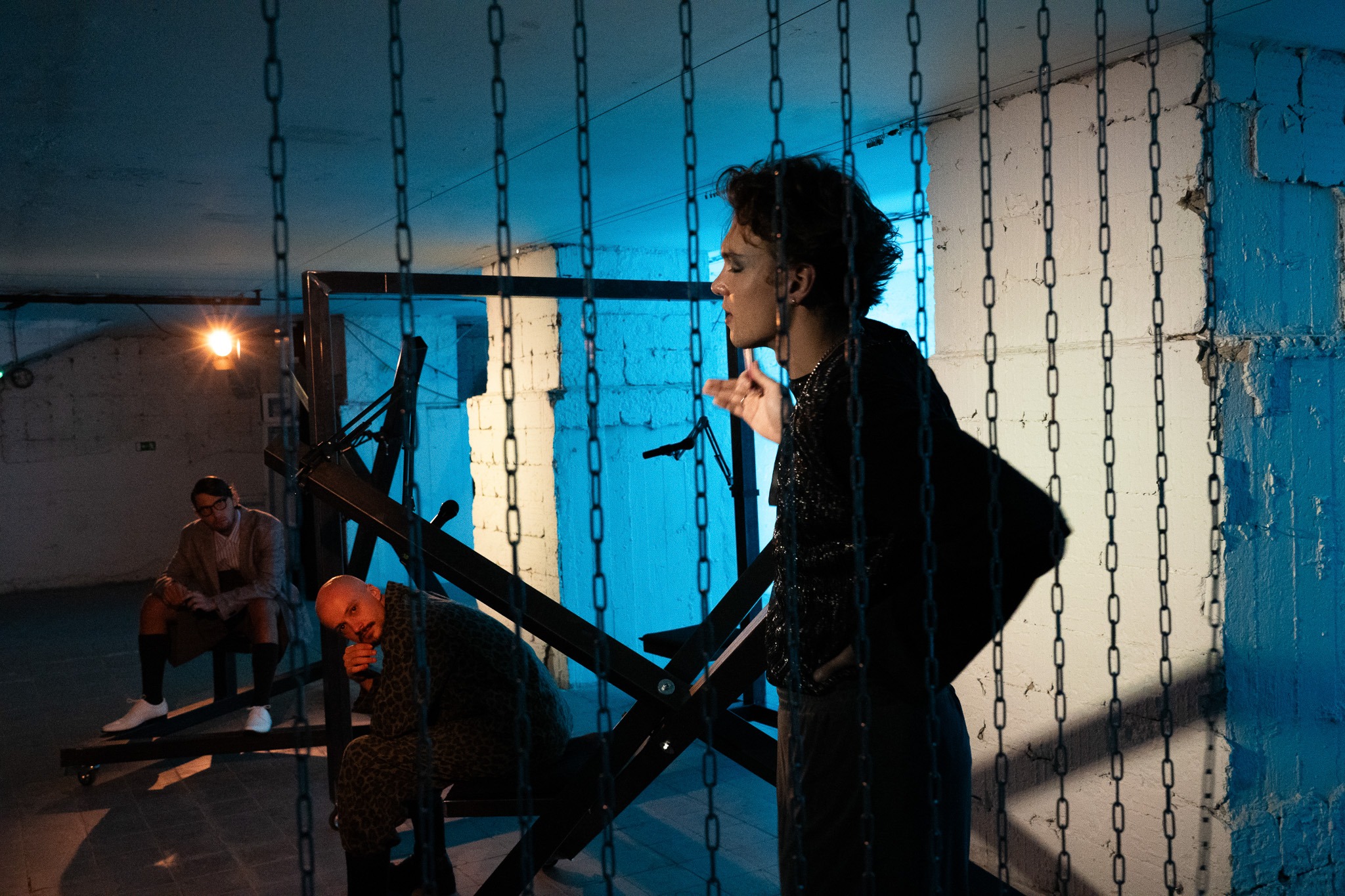 Muž stojí za závesom z reťazí, zatiaľ čo dvaja ďalší chlapi sedia v pozadí v tmavej miestnosti osvetlenej modrým svetlom.