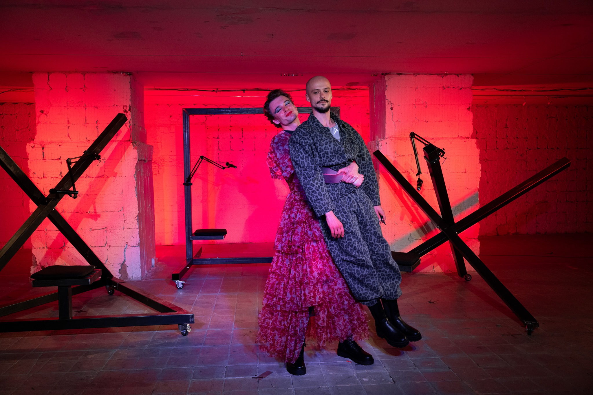 Muž v červených ženských šatách s úsmevom objíma muža v kostýme pripomínajúcom vojenskú uniformu. Druhý muž sa neusmieva.