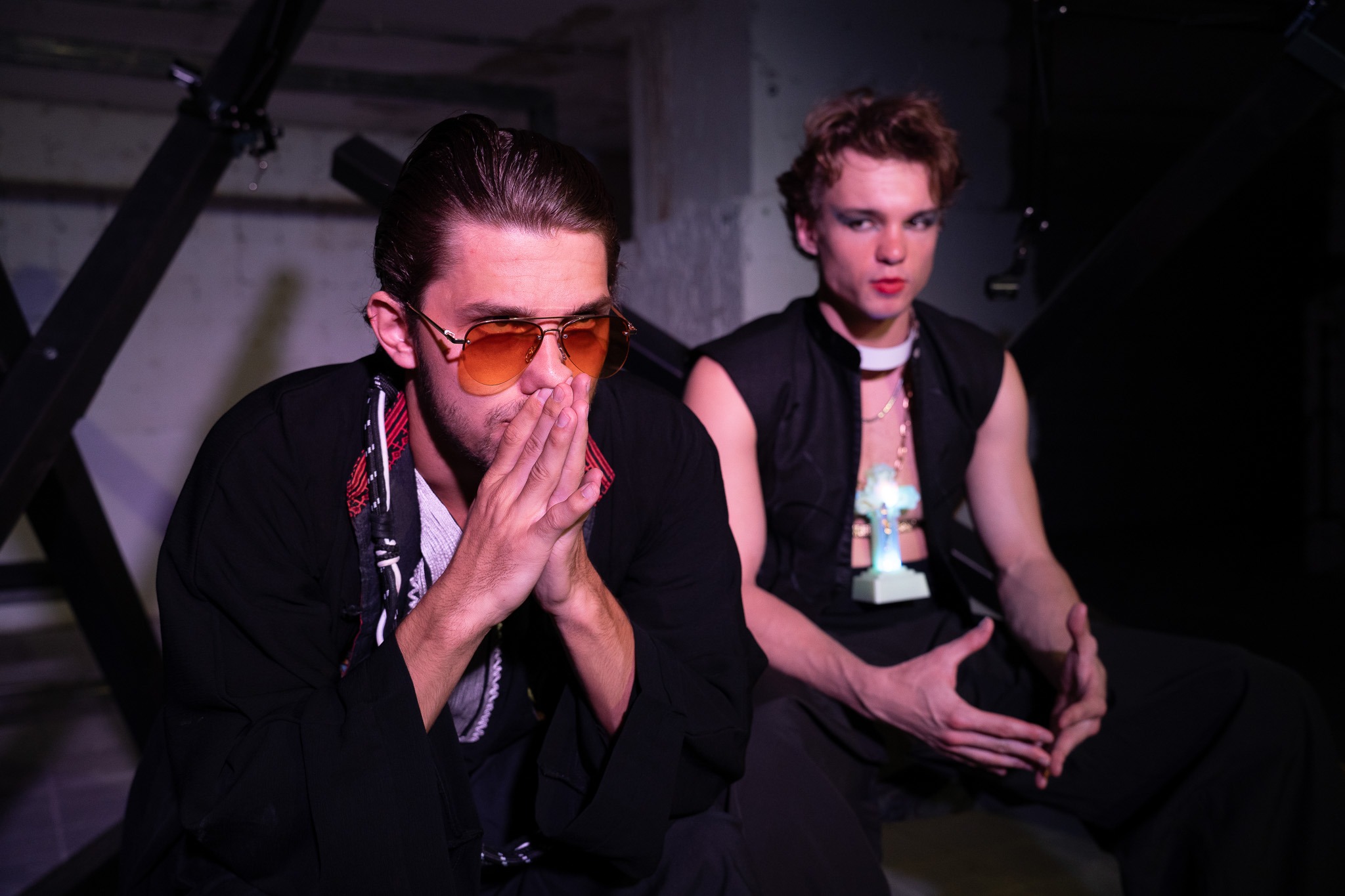 Dvaja muži sedia vedľa seba, jeden má slnečné okuliare a spojené ruky si tisne k ústam, druhý má na tvári výrazný make-up a na krku svietiaci kríž.