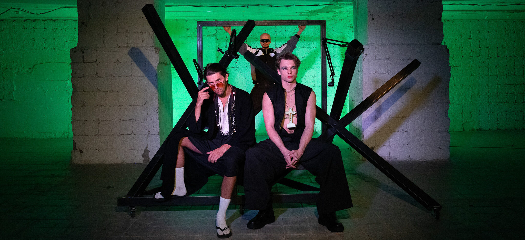 Dvaja muži sedia pred kovovou konštrukciou, tretia postava stojí v pozadí,s rukami nad hlavou sa opiera o konštrukciu, osvetlenie je zelené.