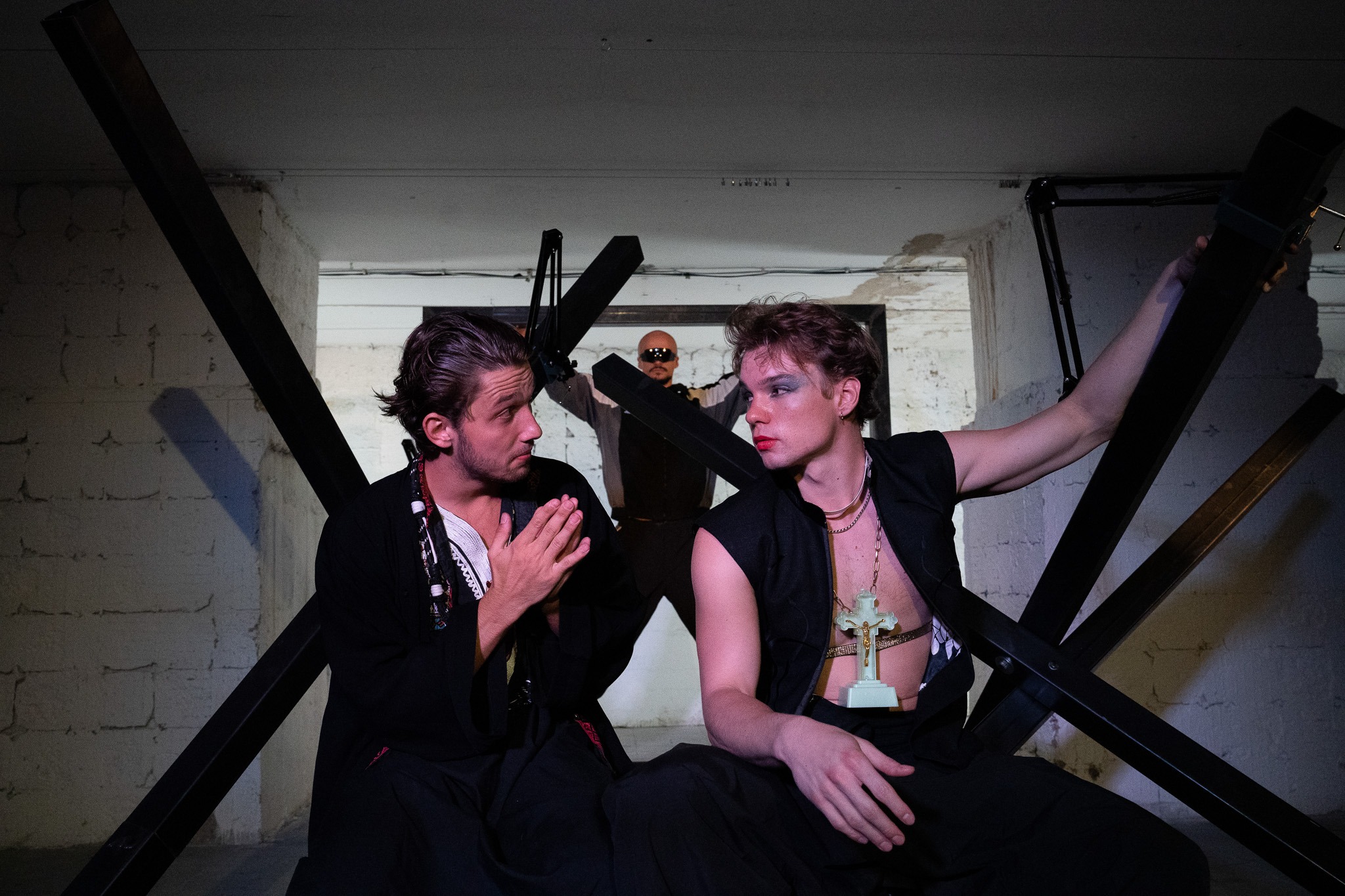 Dvaja muži sedia pred kovovou konštrukciou,pozerajú na seba, tretia postava stojí v pozadí s roztiahnutými rukami nad hlavou.