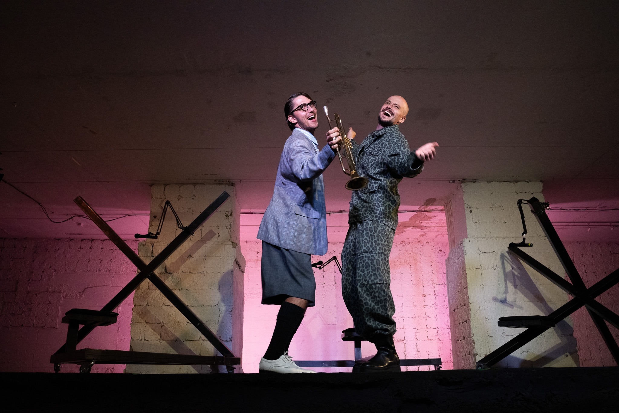 Dvaja muži sa smejú a tancujú, pričom jeden z nich drží trúbku. Obaja stoja pred kovovými konštrukciami na minimalistickej scéne.