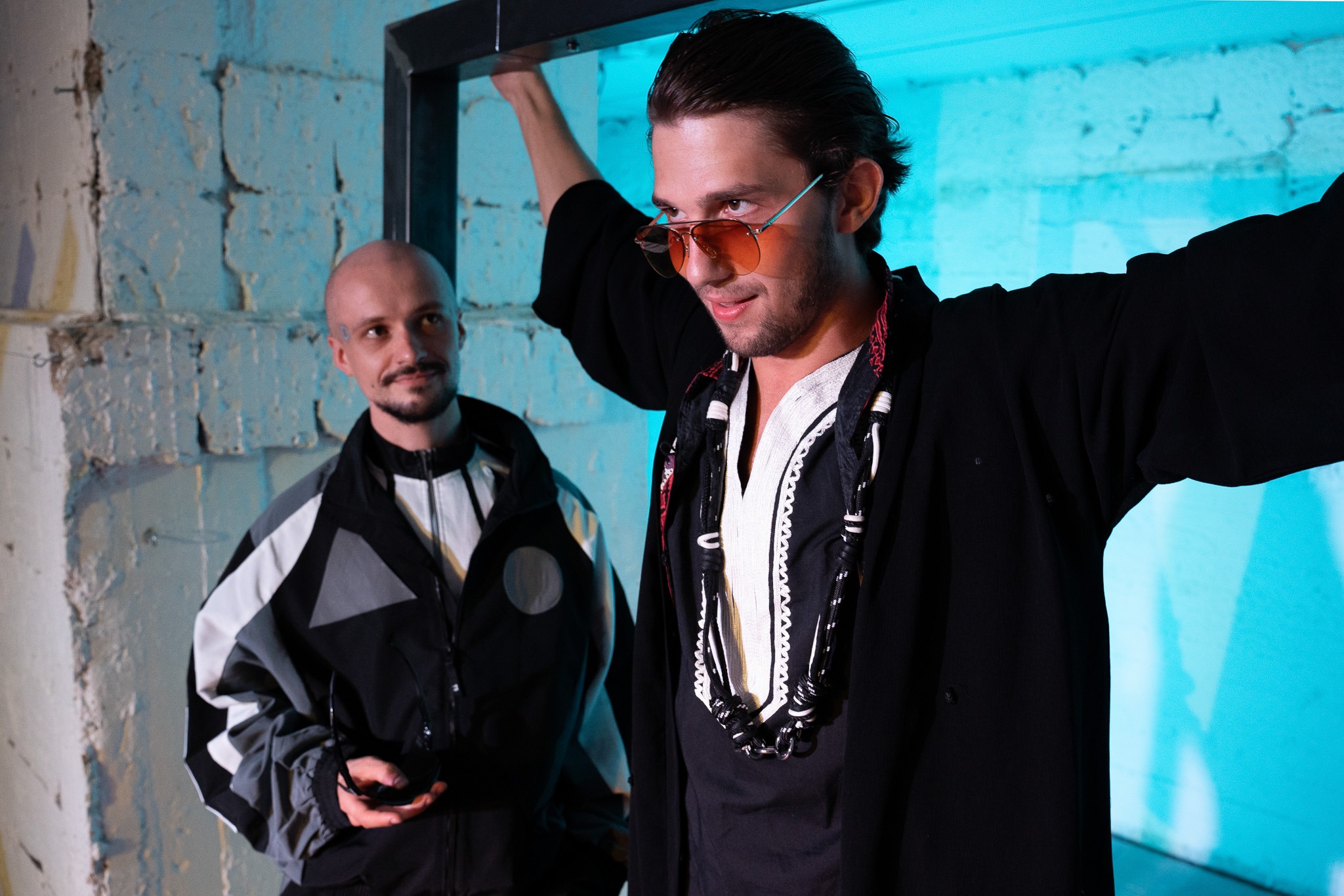 Dvaja muži, jeden v v tmavom oblečení s korálkami na krku a druhý v športovej bunde, stoja vedľa seba na osvetlenej scéne, pričom muž v popredí sa rukami nad hlavou opiera o konštrukciu.