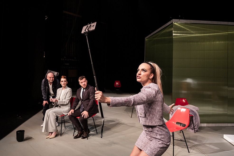 Mladá žena s výrazným make-upom v expresívnej póze natáča selfie video na telefón na selfie paličke. V pozadí ju pozorujú traja ľudia.