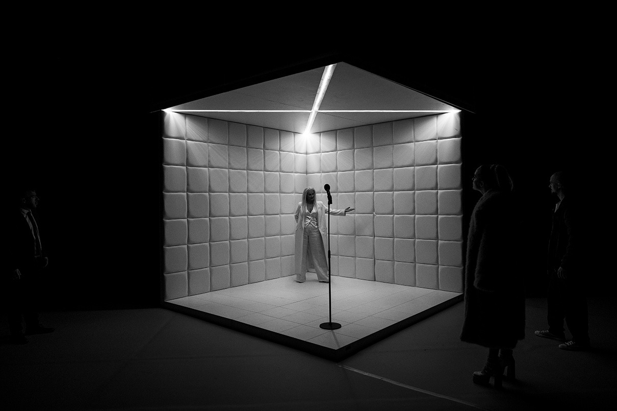 Postava s dlhými blond vlasmi v bielom obleku stojí pred mikrofónom uprostred bielej miestnosti s bielymi mäkkými stenami.