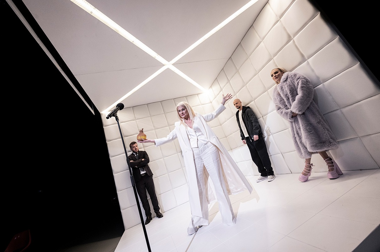 Postava s dlhými blond vlasmi v bielom obleku stojí pred mikrofónom uprostred bielej miestnosti s bielymi mäkkými stenami a drží v ruke maketu cibule. Tri postavy v pozadí ju pozorujú.