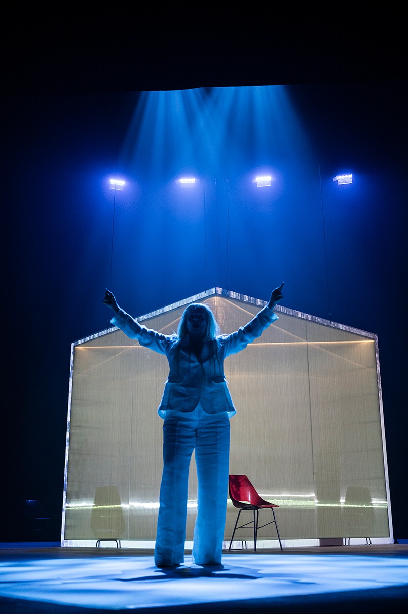 Postava s dlhými blond vlasmi v bielom obleku stojí s rukami natiahnutými do výšky na pódiu s výrazným modrým svietením. V pozadí je miestnosť v tvare kocky a červená stolička.