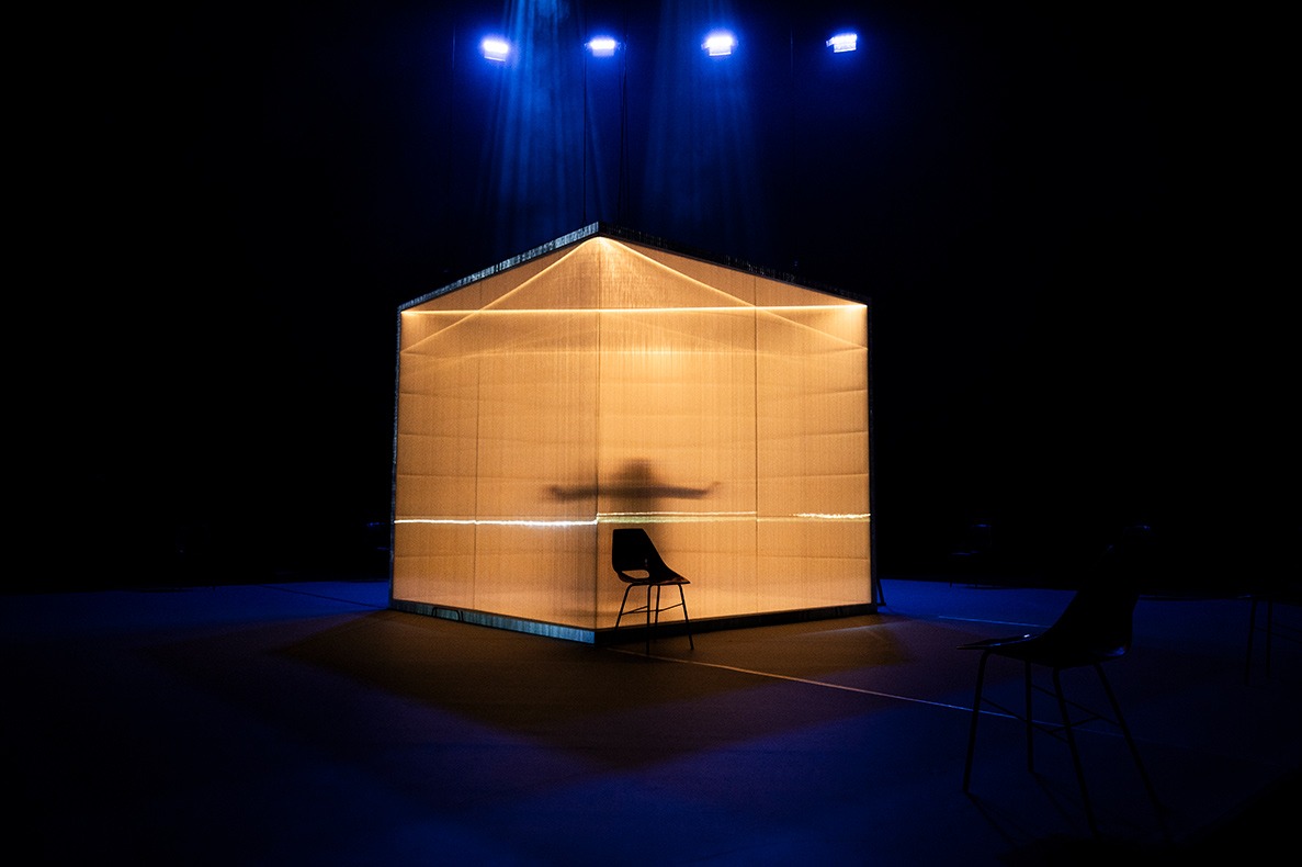 Na pódiu nasvietenom na modro je miestnosť v tvare kocky s priehľadnými plastovými stenami. Vo vnútri vidno ľudskú siluetu s rozpaženými rukami a oranžové svetlo.