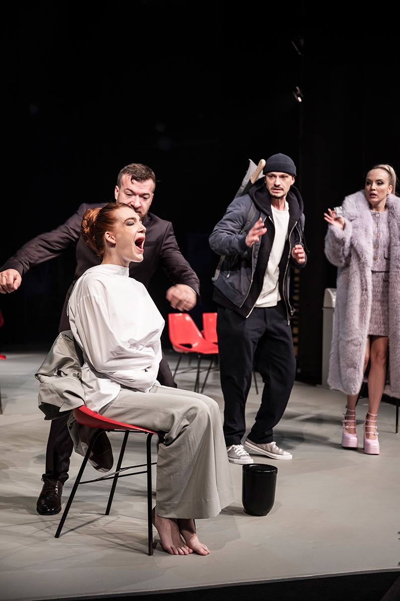 Bosá žena v zvieracej kazajke sedí na stoličke a kričí. Beží ku nej muž v obleku. Ďalší muž a žena ich v pozadí pozorujú.