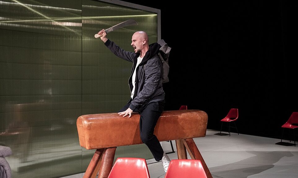 Mladý holohlavý muž sedí v expresívnej póze s rozhnevaným výrazom na gymnastickom koňovi. Zaháňa sa mačetou, ktorú drží v ruke.