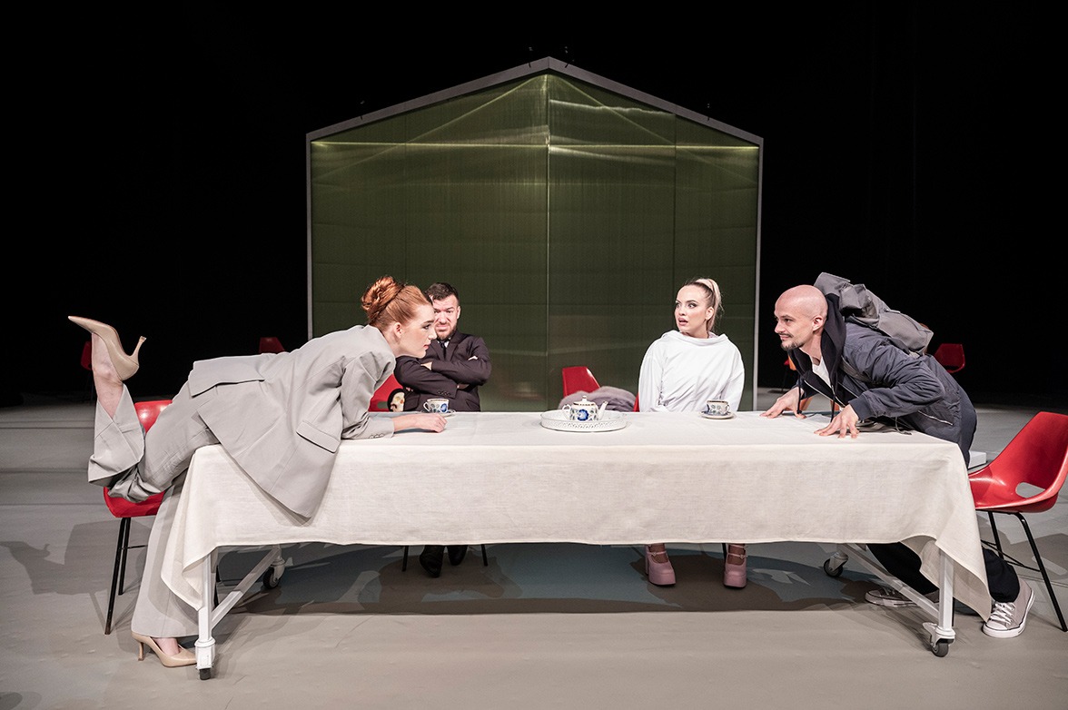 Žena v šedom kostýme sa nakláňa ponad dlhý stôl s bielym obrusom. Oproti nej sa podobne nakľáňa holohlavý mladý muž. Za stolo sedia muž v obleku a žena s výrazným make-upom v zvieracej kazajke.